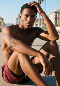 Ing nélküli fiatal férfi bordó rövidnadrágban a járdán ülve, egyik karját a térdén nyugtatja, miközben a haját érinti, mögötte épületek és pálmafák láthatók.