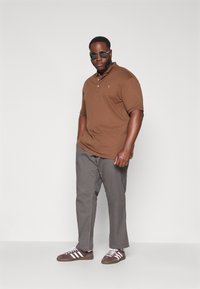 Bruine polo, grijze broek en bruine schoenen met witte strepen. De polo heeft een kraag en twee knopen, gemaakt van zachte stof.