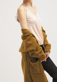 Dlouhý, oversized kardigan v hořčicově žluté barvě, vyrobený z měkkého materiálu, nošený přes světlý růžový top s tenkými ramínky; má velké kapsy a volný střih.