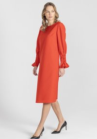 Czerwona sukienka midi z okrągłym dekoltem, długimi bufiastymi rękawami z marszczeniami i gładką fakturą, zestawiona z czarnymi szpiczastymi szpilkami.