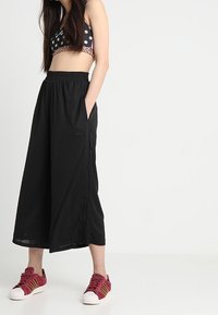 Pantalones culottes negros con una cinturilla elástica, tejido texturizado y bolsillos laterales, combinados con un bralette de lunares y zapatillas rojas.