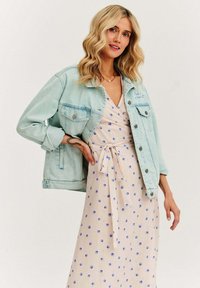 Blondynka w jasnoniebieskiej dżinsowej kurtce na beżowej sukience w niebieski muszelkowy wzór, pozująca z jedną ręką na biodrze przed jednolitą ścianą.