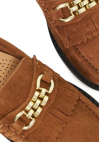 Bruine suede loafers met gouden kettingdetail en franjes op zwarte zolen, weergegeven in close-up.