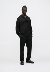 Fekete pamut pulóver gallérral és nagy "HUGO" logóval, hozzáillő fekete nadrággal, hasonló logóval, fekete cipővel viselve.