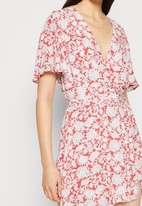 Rode bloemenjurk met een diepe V-hals, korte fladdermouwen en een wikkeldesign met witte botanische patronen. Zachte stoftextuur.