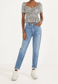 Kobieta w bluzce z rękawami w kształcie bufki w panterkę, w niebieskich dżinsach z wysokim stanem z podwiniętymi nogawkami oraz białych trampkach, stoi z jedną ręką w kieszeni.