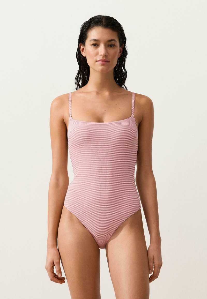 Femme aux cheveux mouillés portant un maillot de bain une pièce rayé rose et blanc avec de fines bretelles, sur un fond clair uni.