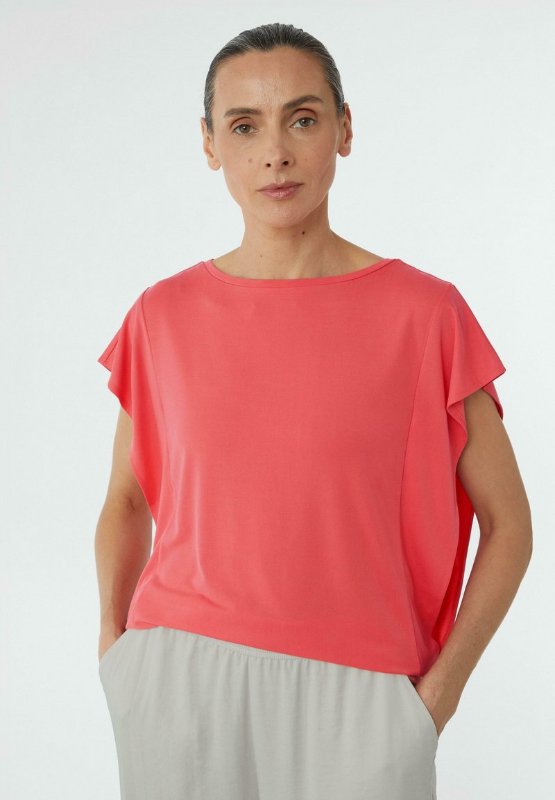 Koronkowa bluzka z krótkim rękawem w kolorze koralowym o luźnym kroju, z dekoltem w łódkę oraz miękkiego materiału. W komplecie z jasnoszarymi, zwiewnymi spodniami.