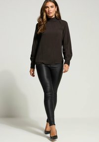 Musta pitkähihainen paita, jossa on korkea kaulus ja laskostettu yksityiskohta, yhdistettynä tiukkoihin mustiin nahkahousuihin ja mustiin kärkitossuihin.