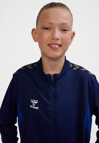 Marinblå sportjacka med hel dragkedja, andningsbar textur och kontrasterande blå sidopaneler. Har mönstrade svarta axeldetaljer och logotyp.