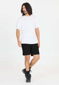 Vit bomull T-shirt, svarta shorts med dragkedjor och svarta sportskor. Modellen poserar stående med ett neutralt uttryck.