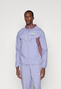 Άνδρας φορώντας ένα ανοιχτό μοβ και μωβ μπουφάν Nike με ταιριαστά ανοιχτό μοβ παντελόνια, μπροστά σε απλή ανοιχτή φόντο.