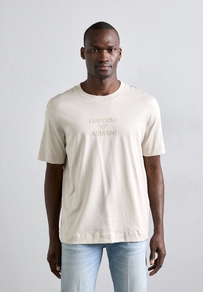 Beige bavlněné tričko s vyraženým logem "Emporio Armani" na přední straně. Krátké rukávy a kulatý výstřih, nošené se světle modrými džíny.