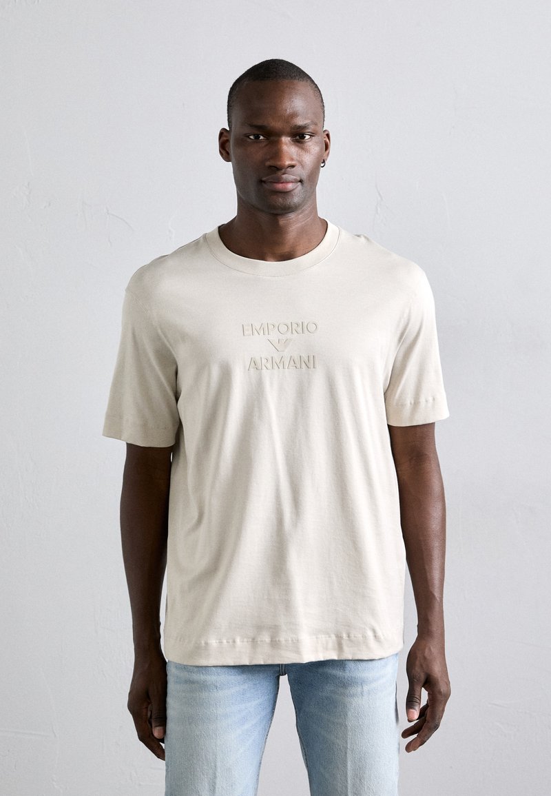 Μπεζ βαμβακερό T-shirt με ανάγλυφο λογότυπο "Emporio Armani" στο μπροστινό μέρος. Έχει κοντά μανίκια και στρογγυλή λαιμόκοψη, συνδυασμένο με ανοιχτόχρωμα τζιν.
