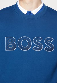 Blå sweatshirt med ribbad rund halsringning, prydd med en stor broderad "BOSS" logotyp i kontrasterande vitt med en texturerad yta.