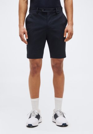 Mees, kes kannab musta kohandatud lühikesi pükse, valgeid resoliidseid sokke ja valgeid tossusid mustade detailidega, seisab tavalise tausta ees.