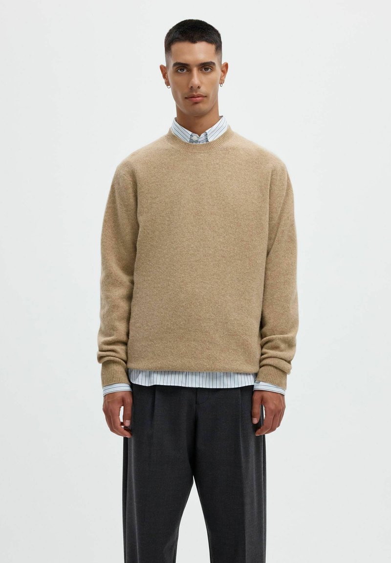 Jeune homme portant un pull beige par-dessus une chemise rayée avec un pantalon sombre, debout devant un fond blanc uni.