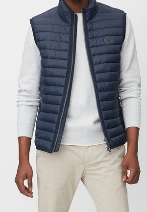 Mand iført en marineblå quiltet vest over en lysegrå sweater og beige bukser, stående med armene afslappet langs siden.