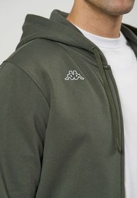 Mann trägt eine olivgrüne Kapuzenjacke mit Reißverschluss und weißem Kappa-Logo auf der Brust sowie ein weißes Rundhals-T-Shirt darunter.
