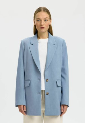 Vrouw draagt een lichtblauwe oversized blazer met twee knopen over een wit overhemd en beige broek, staand tegen een effen achtergrond.