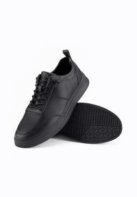Baskets noires en cuir avec tige texturée, semelle en caoutchouc plate, motif de grip hexagonal et design classique à lacets. Onglet de tirage en accent sur le talon.