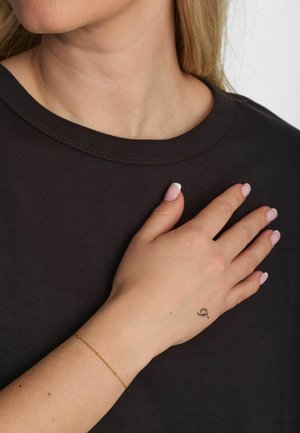 Czarny bawełniany t-shirt z okrągłym dekoltem; ręka spoczywająca na klatce piersiowej z delikatną złotą bransoletką i małym tatuażem na nadgarstku.