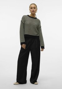 Femme se tenant debout, portant un pull à rayures noires et blanches, un pantalon noir à jambes larges et des chaussures plates beiges, sur un fond blanc uni.