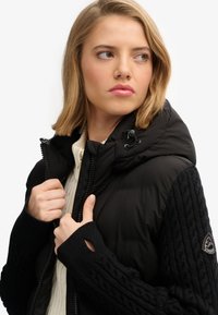 Zwarte puffer vest met een kabelbreipatroon op de mouwen, dubbele ritsen en een capuchon met trekkoord. Bevat een logo-opdruk op de linkerarm.