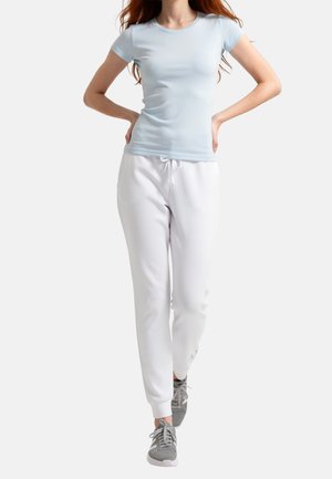Donna con t-shirt aderente azzurro chiaro, pantaloni della tuta bianchi con coulisse e sneakers grigie, in piedi con le mani sui fianchi su sfondo uniforme.