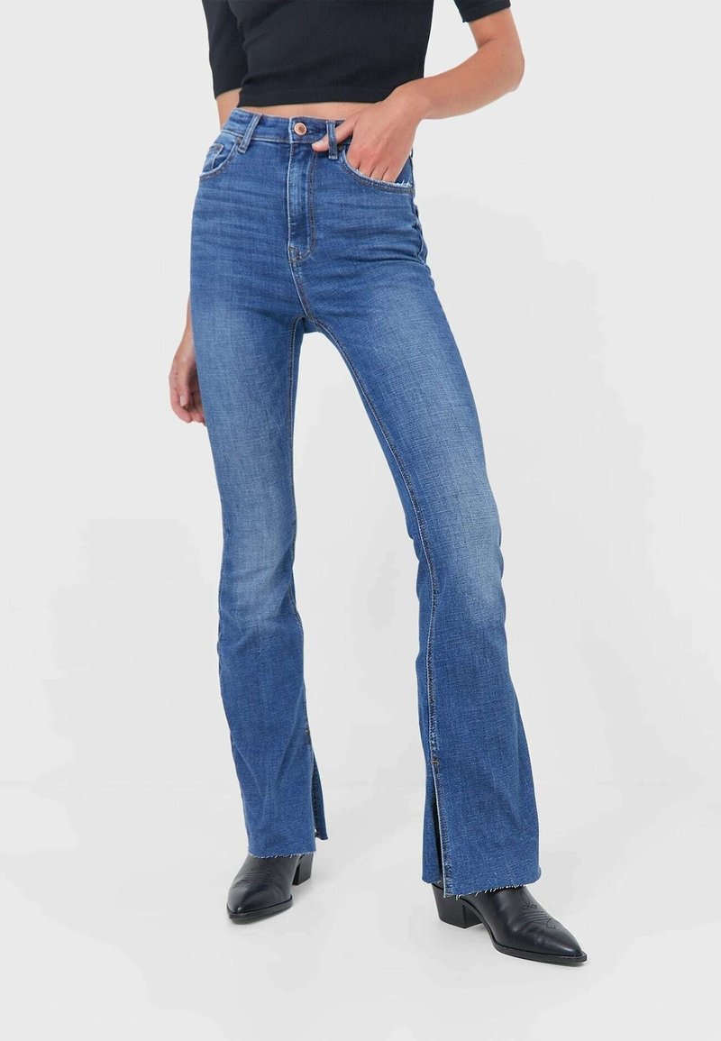Femme portant un jean en denim bleu taille haute avec des fentes sur le devant et des bottines noires, main dans la poche, sur un fond uni.