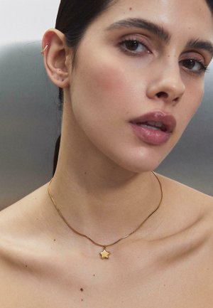 Kobieta z gładką skórą nosząca cienki złoty naszyjnik z zawieszką w kształcie gwiazdy oraz małą złotą obręczkę na lewym uchu.