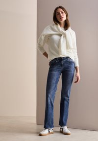 Suéter blanco texturizado colgado alrededor del cuello, combinado con jeans de denim azul y zapatillas blancas con detalles en negro. Conjunto simple y casual.