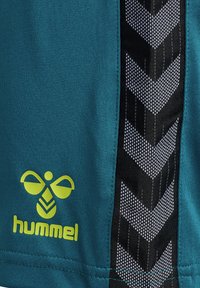 Turkosa sportshorts med svart sidopanel som har ett geometriskt mönster och reflektiva detaljer. Gul "hummel"-logga framtill.