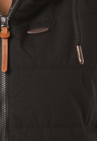 Schwarze Jacke mit Reißverschluss und glatter Textur, mit einem braunen Lederdetail und metallischen Akzenten. Das Design umfasst sichtbare Nähte und eine Kapuze.