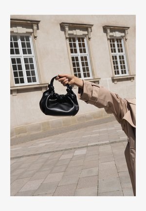 Bras portant un trench beige tenant un petit sac à main en cuir noir devant un bâtiment beige avec trois fenêtres.