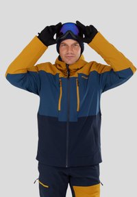 Veste de ski en bleu, navy et moutarde avec une capuche, des poches zippées et des poignets ajustables ; assortie avec des lunettes de ski tenues par une main gantée.