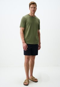 Oliwkowy t-shirt z krótkim rękawem, wykonany z miękkiej bawełny, w połączeniu z ciemnogranatowymi szortami. Model nosi beżowe sandały z krzyżowym paskiem.