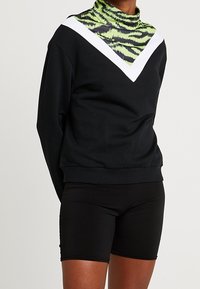 Persona vestida con pantalones cortos negros y un sudadera negra con un patrón de chevron blanco y rayas de tigre verde neón en el cuello y el pecho.