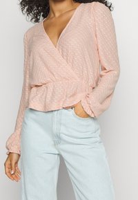 Rosa texturerad omslagstopp med långa ärmar och djup V-ringning, ihopparad med ljusblå högmidjade jeans. Tyget har ett subtilt mönster.