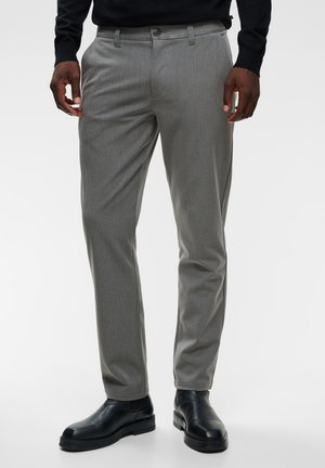 Homme portant un pantalon gris tailleur avec poches latérales et des chaussures de ville en cuir noir sur un fond uni.