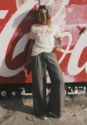 Biała koszulka z czerwonymi akcentami, spodnie w szerokie paski, czarno-białe trampki w kratę, trzymająca czerwoną puszkę Coca-Coli, na tle vintage.