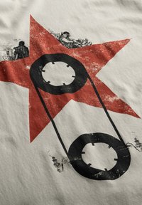 T-shirt met een crèmekleurige achtergrond, voorzien van een grote rode ster en een zwart veiligheidsspeld graphic, versierd met gedetailleerde zwarte illustraties eromheen.
