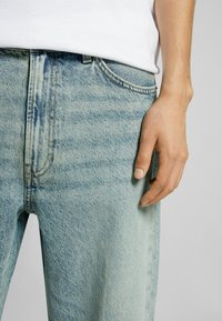 Mano derecha descansando junto al bolsillo de unos vaqueros de lavado ácido azul claro, vistiendo una camisa blanca sobre un fondo liso.