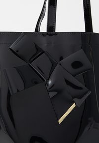 Svart lackläder tote-väska med en strukturerad design, en stor rosett och en logoplatta i guldton. Slät, glansig yta.