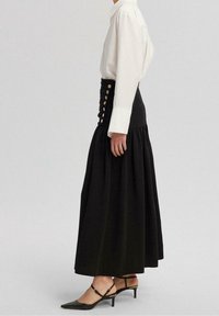 Schwarzer Maxirock mit passgenössigem Taillenbund, goldenen Knöpfen, einem ausgestellten Design und geschichteter Textur; kombiniert mit einem weißen Hemd und schwarzen Heels.