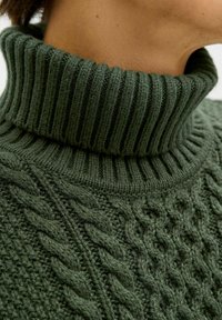Pull en maille verte à col roulé avec un col à côtes et un design en tricot torsadé texturé, mettant en avant des motifs complexes et une douce texture en laine.