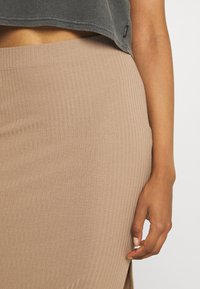 Jupe en maille beige côtelée avec une fente latérale, tissu texturé et coupe ajustée. La main du mannequin repose sur l'ourlet de la jupe.