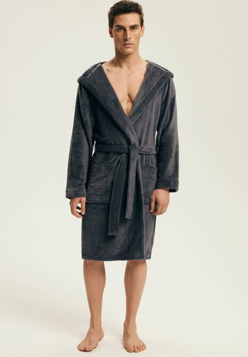 Man standing barefoot wearing a dark gray hooded bathrobe tied at the waist with large front pockets, plain background.