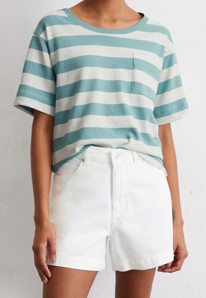 Person in einem türkis-weiß gestreiften Shirt mit Brusttasche und weißen, hoch taillierten Shorts vor einer weißen Wand.