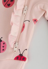Tela rosa claro con estampados de mariquitas rojas, una cremallera blanca y un pequeño detalle de volantes a lo largo de la costura de la cremallera en una prenda.